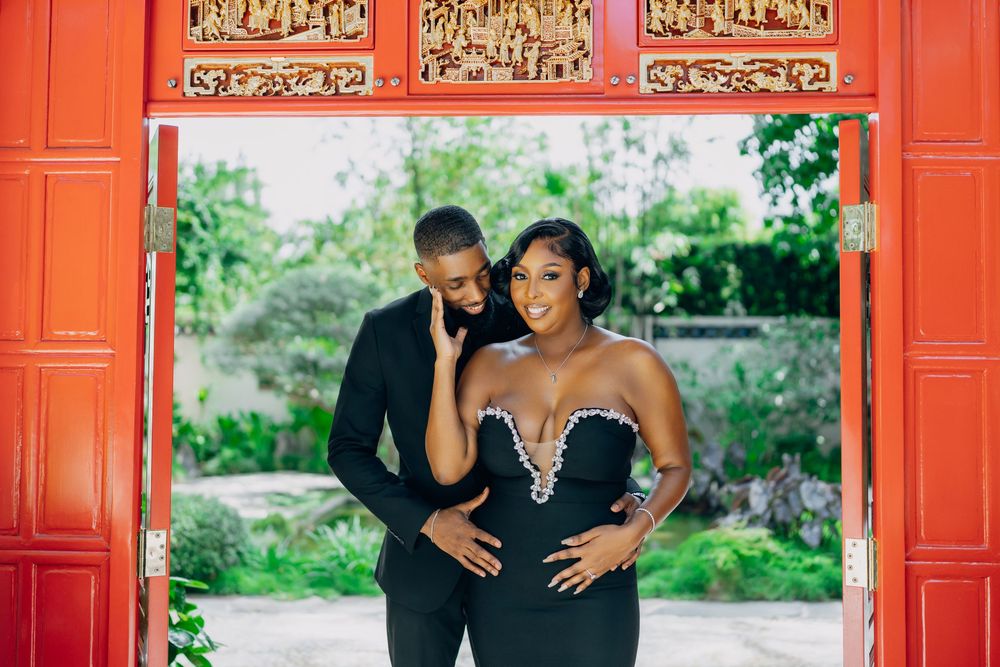 A romantic couple shares a joyful moment together under a red decorative doorway in a garden setting.
