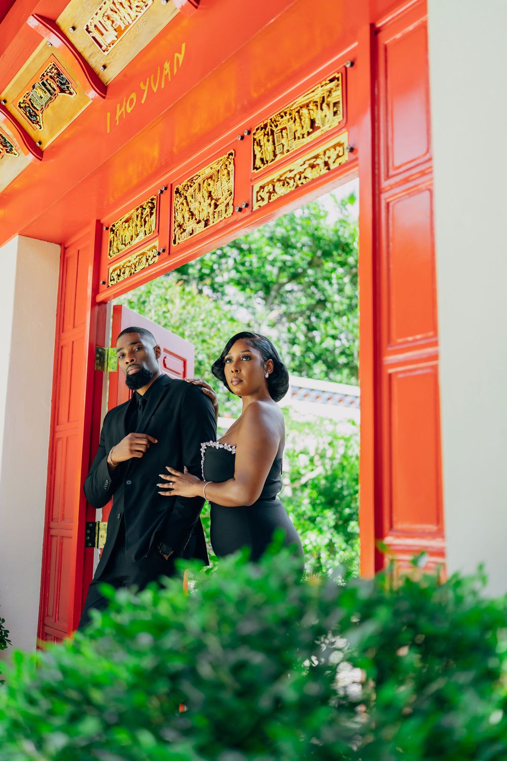 Couple stands beneath bright red temple entrance with ornate gold trim and lush greenery in the foreground.