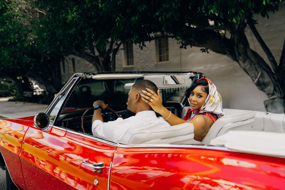 Two people smile and laugh together while sitting in a bright red vintage convertible car parked under trees.
