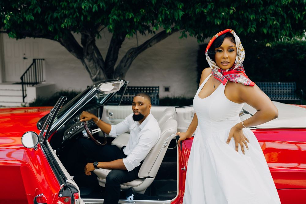 A glamorous couple poses with a classic red convertible car, with the person in white wearing a head scarf.