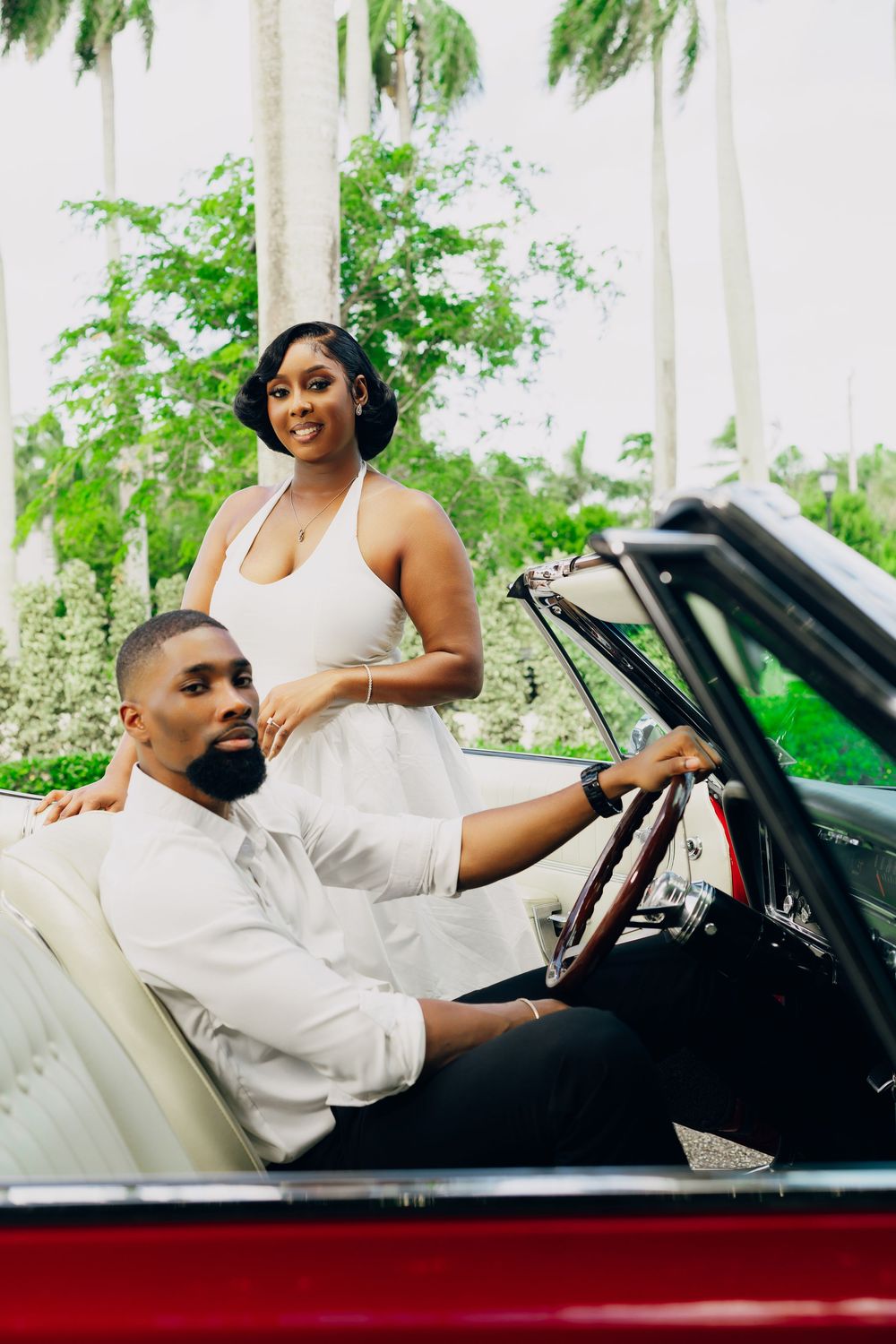Stylish couple poses with classic red convertible car surrounded by palm trees and lush greenery.