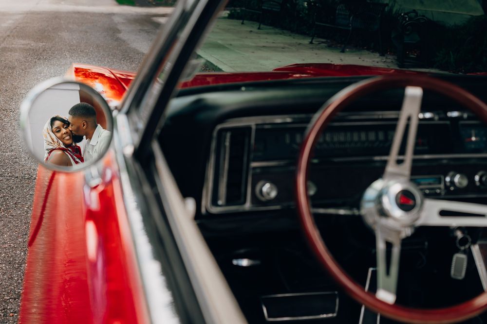 View through side mirror of vintage red car showing classic wooden steering wheel and dashboard interior.
