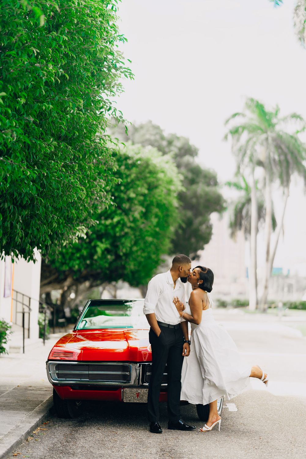 Two people sharing a romantic moment next to a vintage red convertible car on a palm tree-lined street.