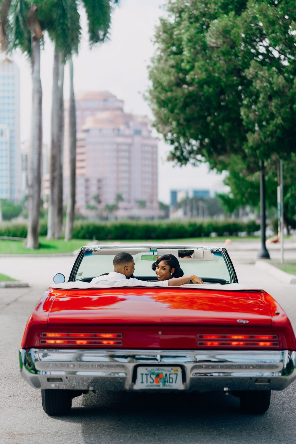 A couple sitting in a vintage red convertible car with palm trees and city buildings in the background.