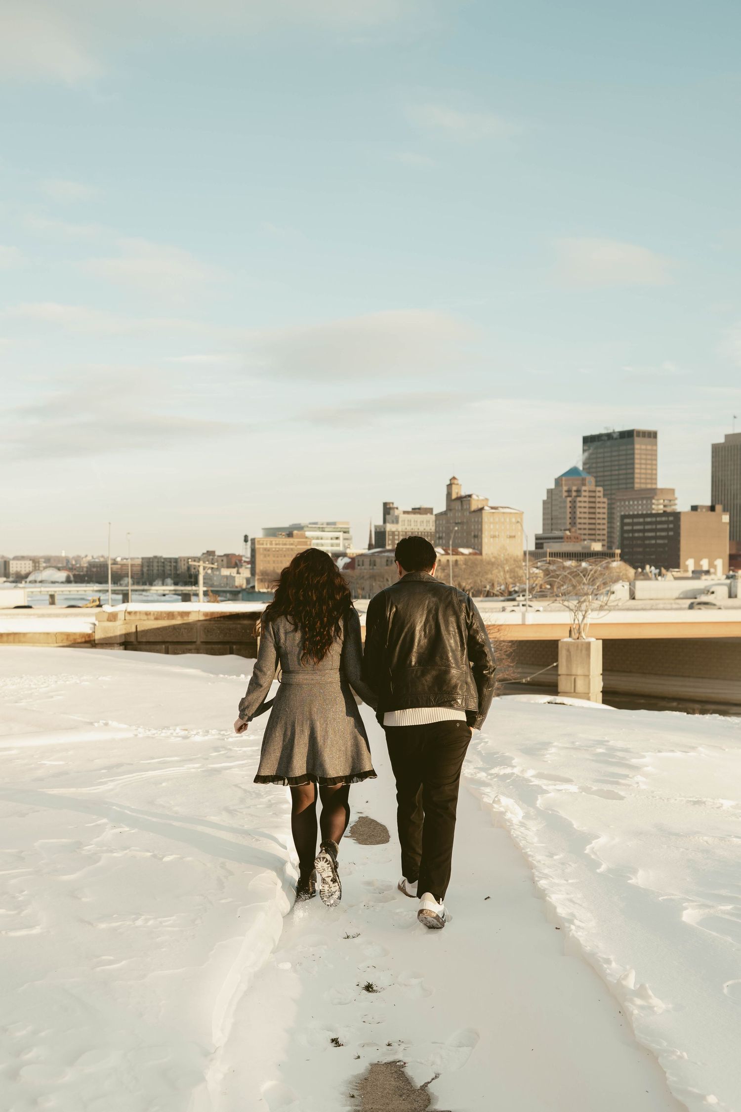 Couple walk together on a snowy path with buildings visible in the winter landscape at Dayton Art Institute during engagement session.