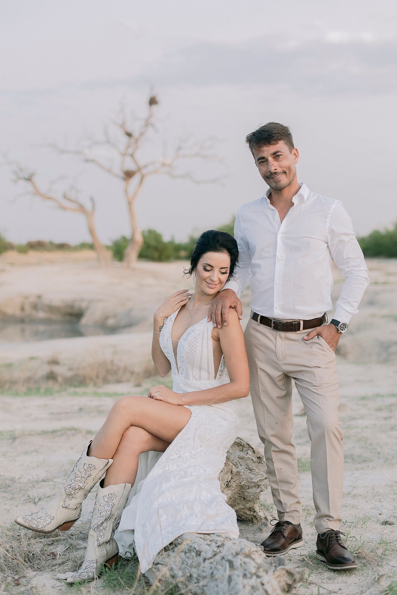 Couple poses on rocky savanna landscape with bare tree in background.