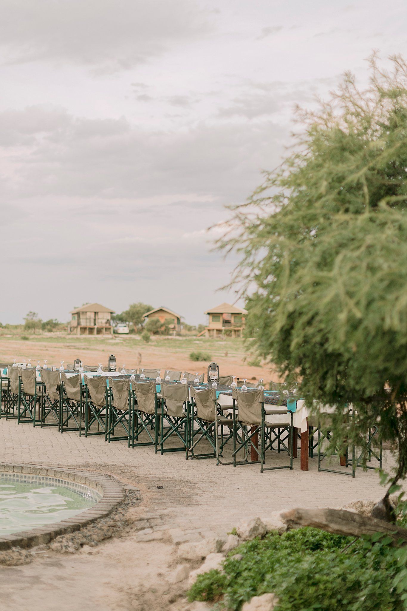 Long outdoor dining table setup with safari chairs beside a pool at African bush lodge.