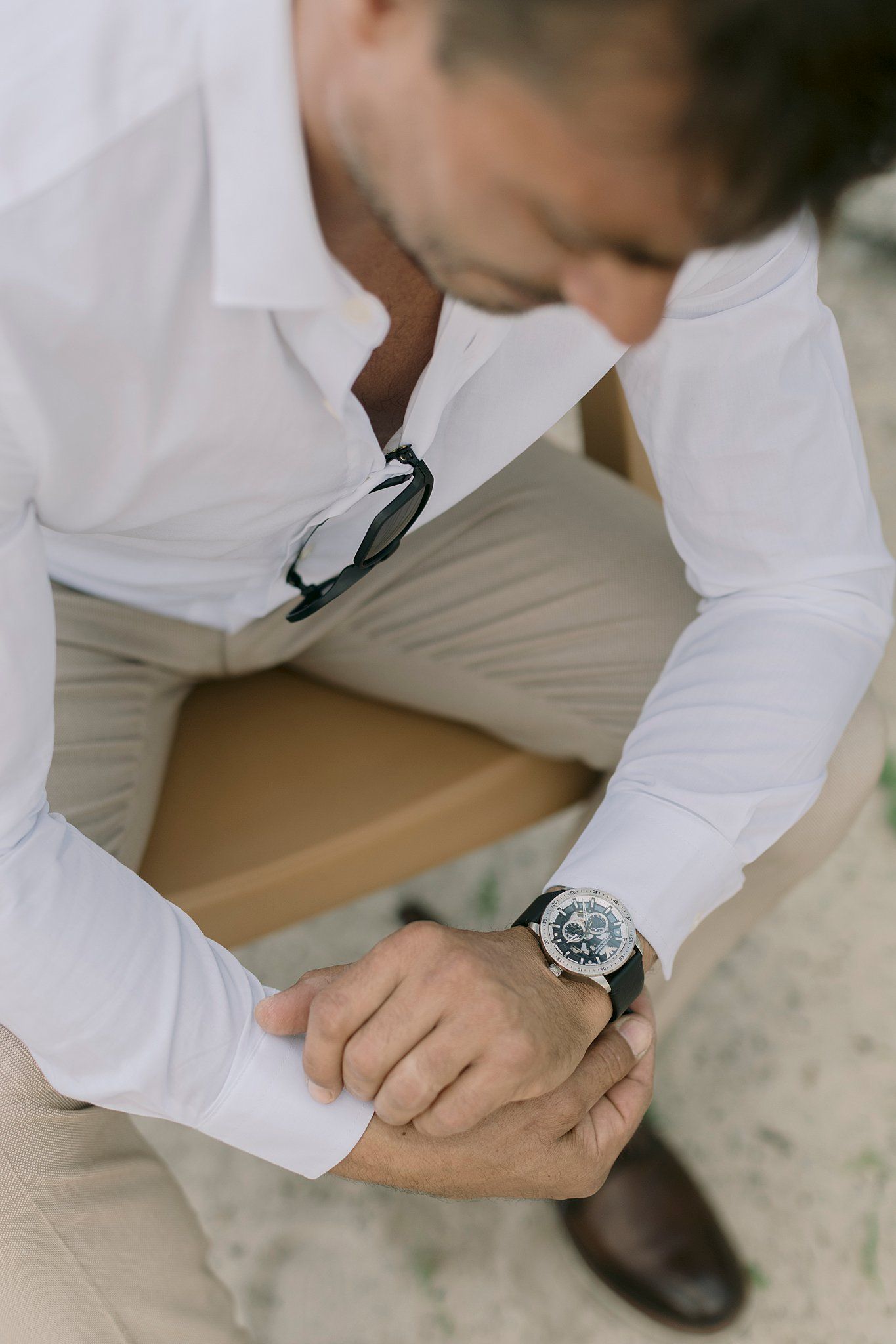 Close-up of a groom in white shirt checking his luxury mechanical wristwatch while seated on a wooden chair.