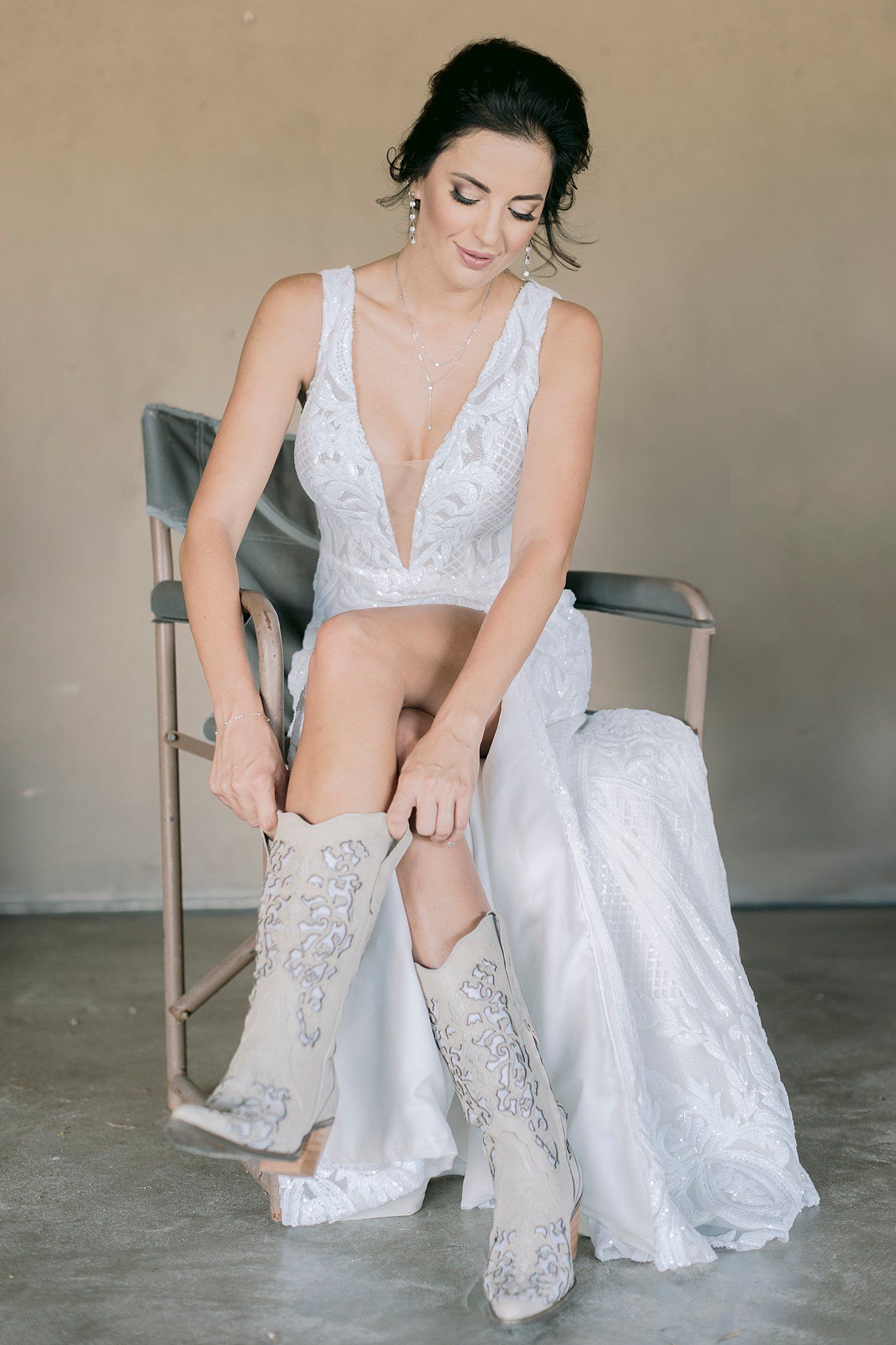 Bride in white lace wedding dress adjusting embroidered cowboy boots while seated.