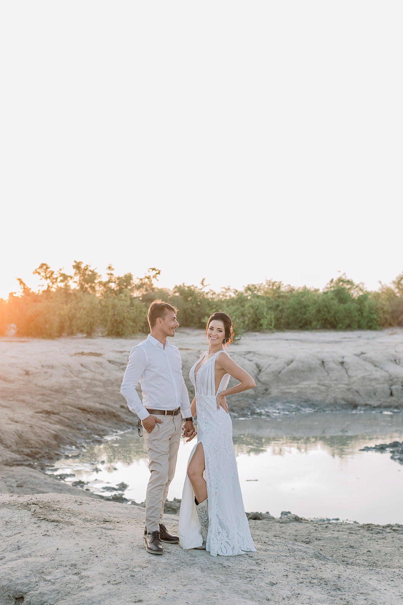 Bride and groom stand by a sunlit sandy riverbank at golden hour sunset.