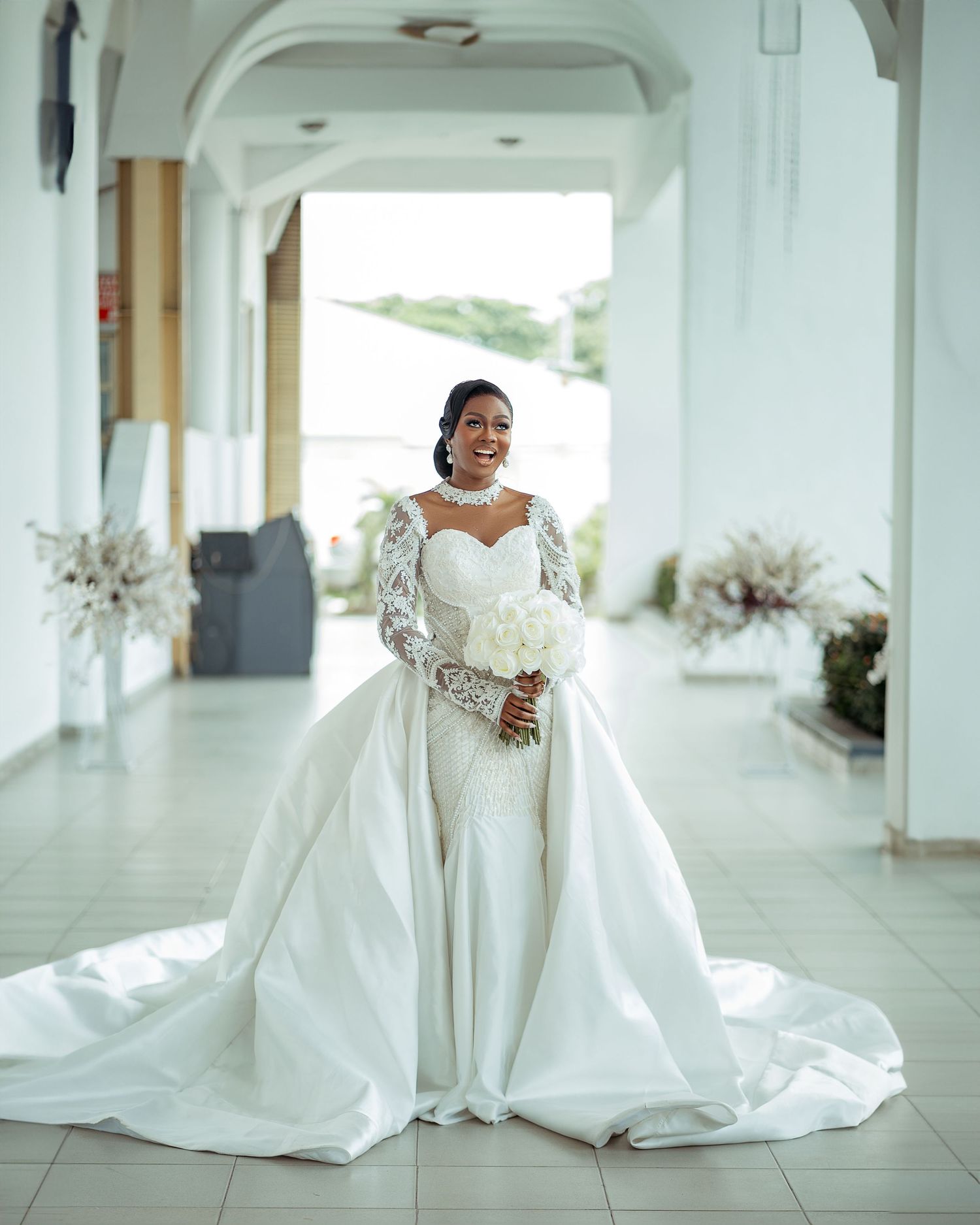 Elegant bride in a stunning white off-shoulder ball gown with long train poses in a bright columned hallway.