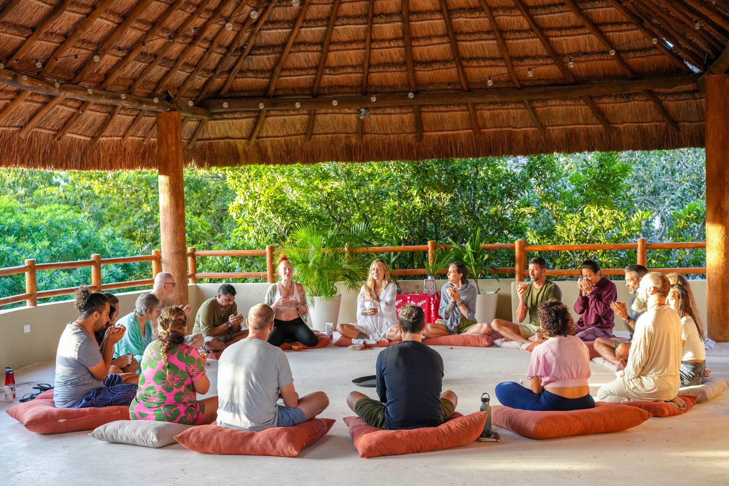 A group gathers for meditation under a thatched-roof pavilion with tropical surroundings.