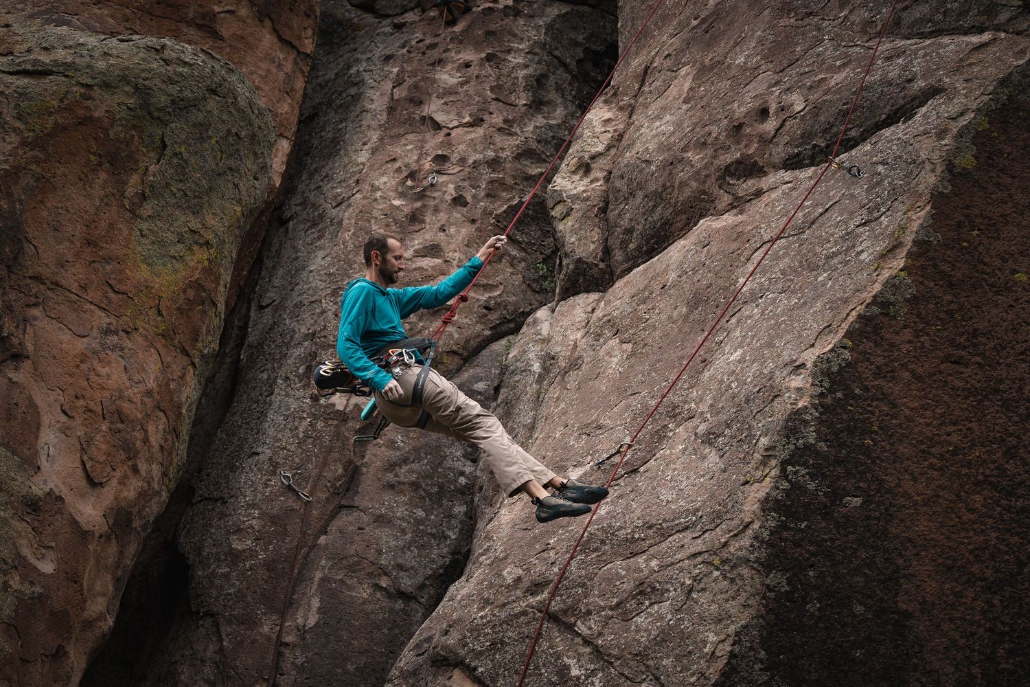 A rock climber in a turquoise jacket scales a steep cliff face using ropes and climbing gear.