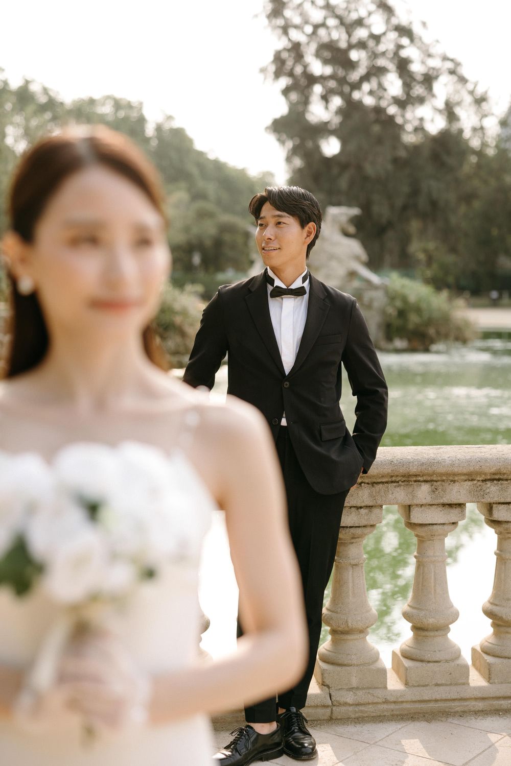 Elegant wedding couple poses on stone balustrade in garden setting with soft natural lighting.