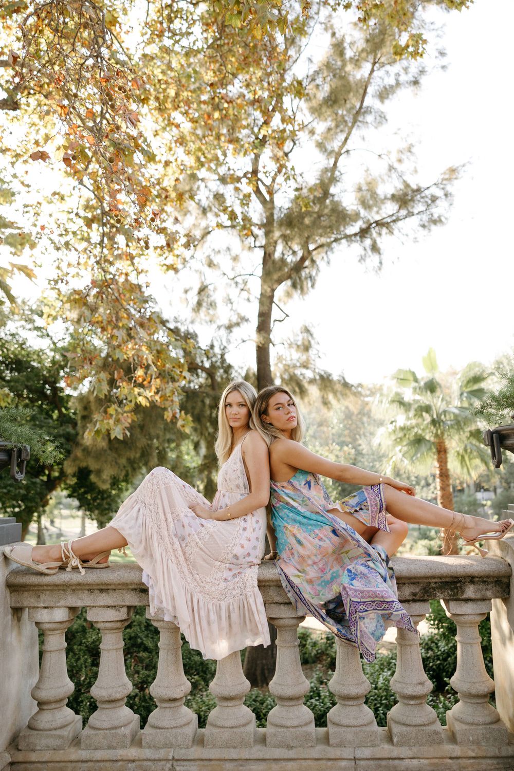 Two friends relax on stone balustrade during golden hour surrounded by autumn trees.