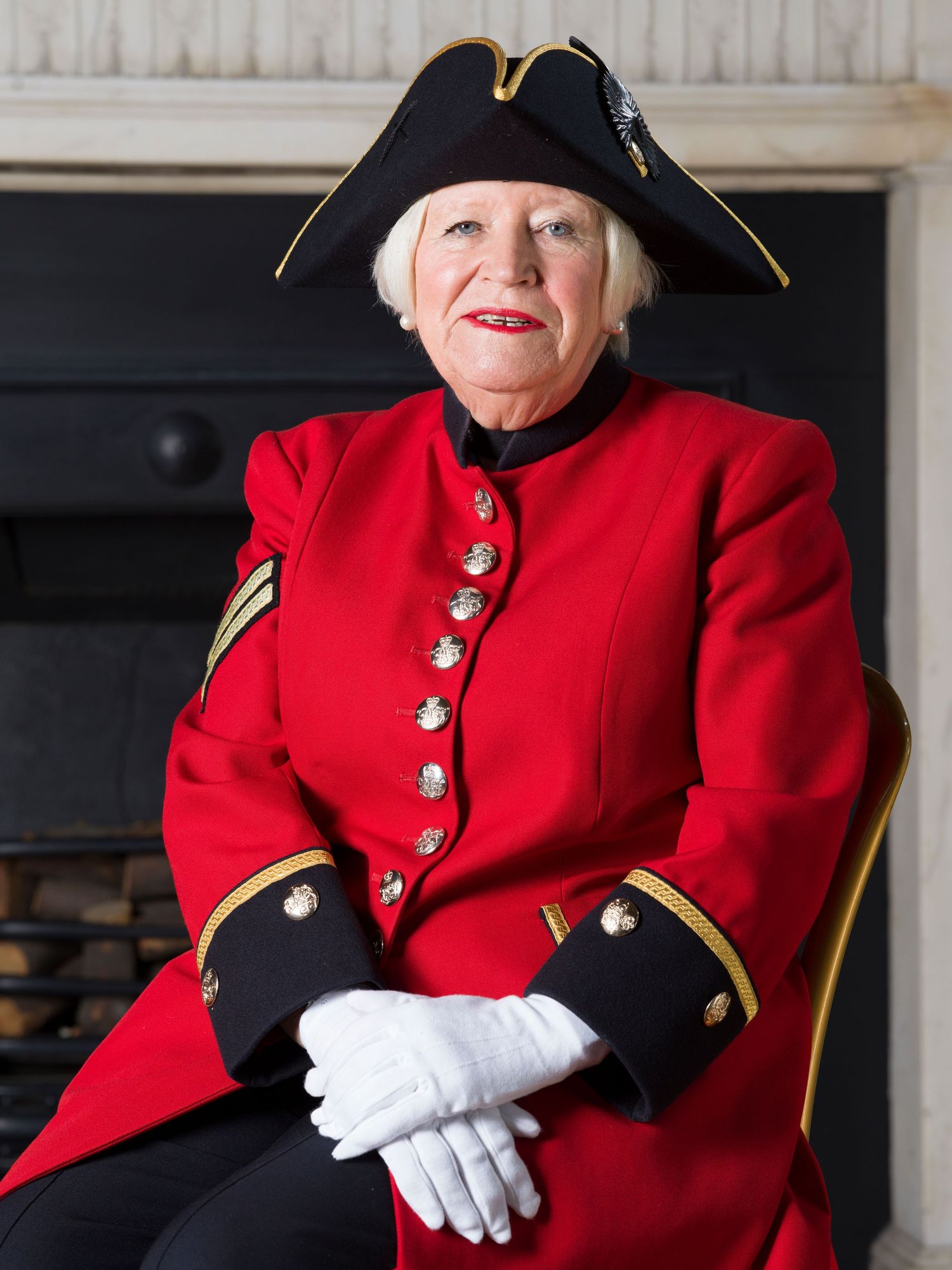 Someone wearing a traditional red and black Chelsea Pensioner uniform with tricorn hat and white gloves sits in formal pose.