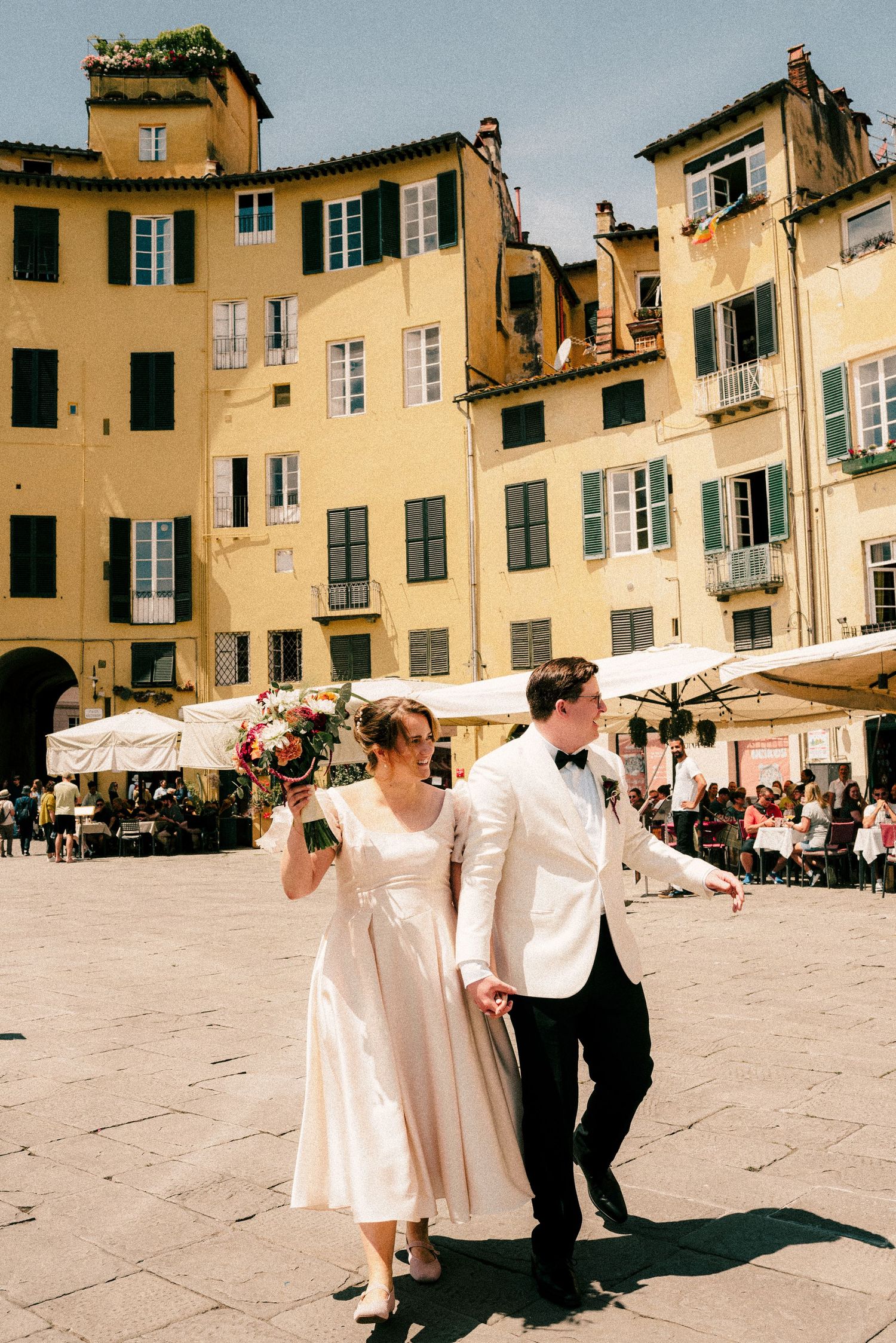 A wedding couple stands in front of historic yellow buildings in an Italian plaza.