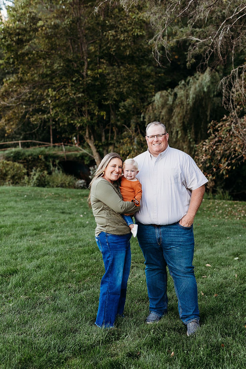 A family poses together on a grassy lawn with trees and foliage in the background during a fall day.