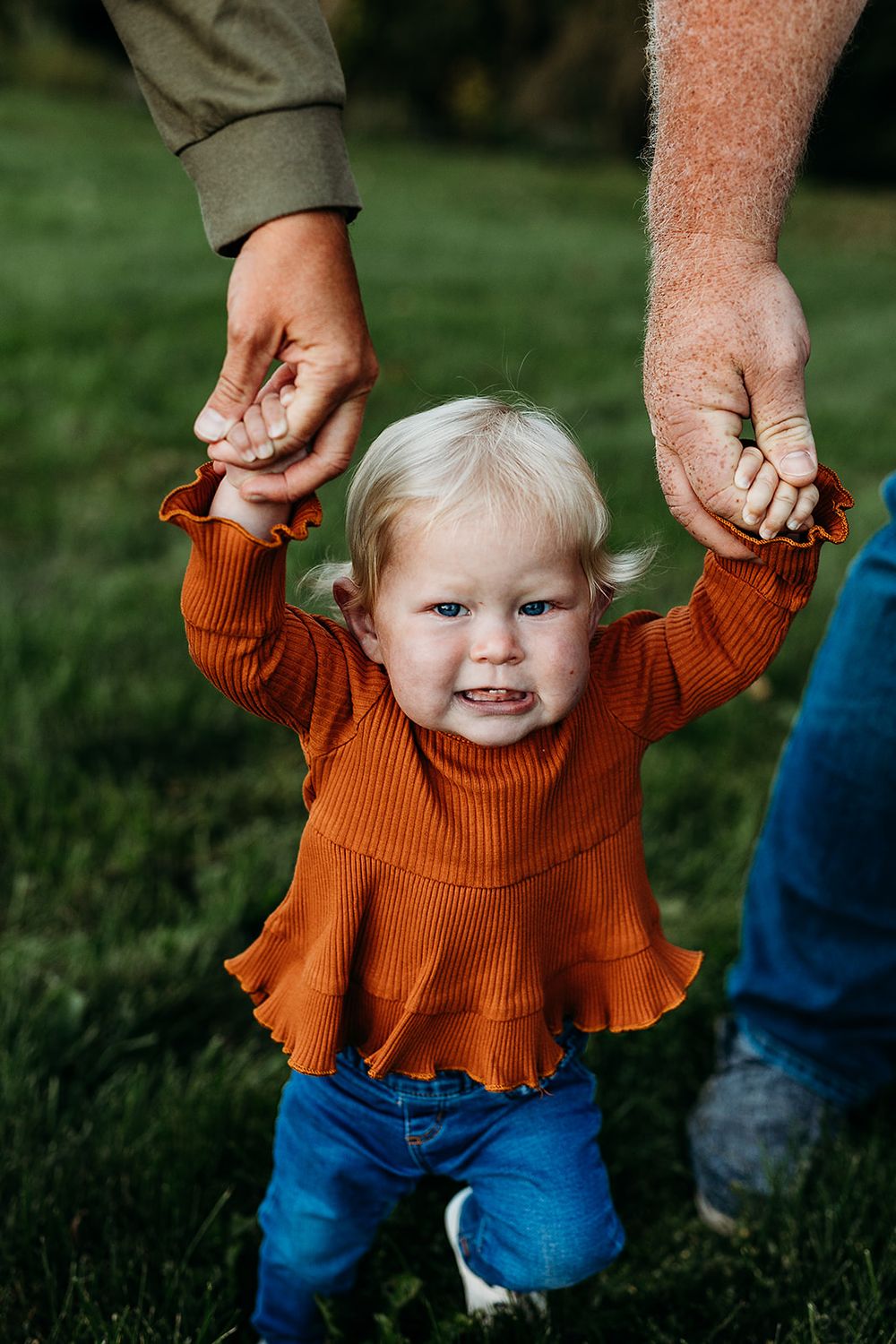 A small child in an orange sweater smiles while being supported by two hands on a grassy lawn.
