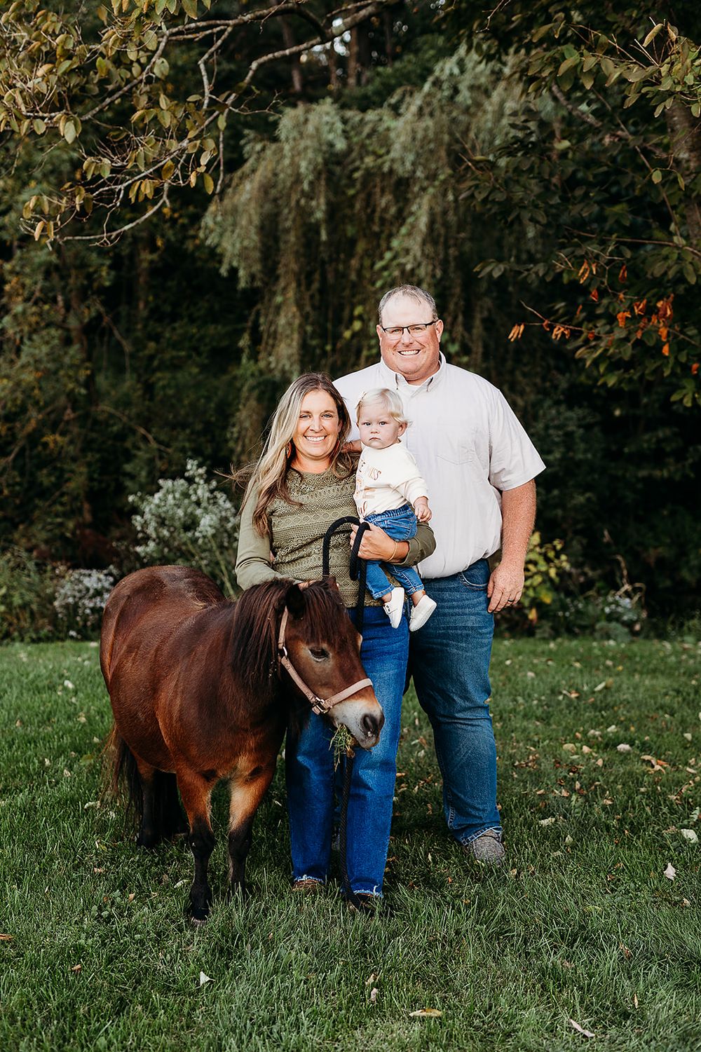 A family poses with their miniature horse in a garden setting surrounded by autumn foliage.