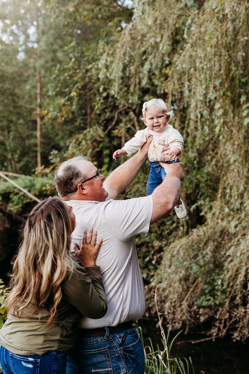 Couple lifts small child joyfully near a weeping willow tree beside a peaceful pond on a sunny day.