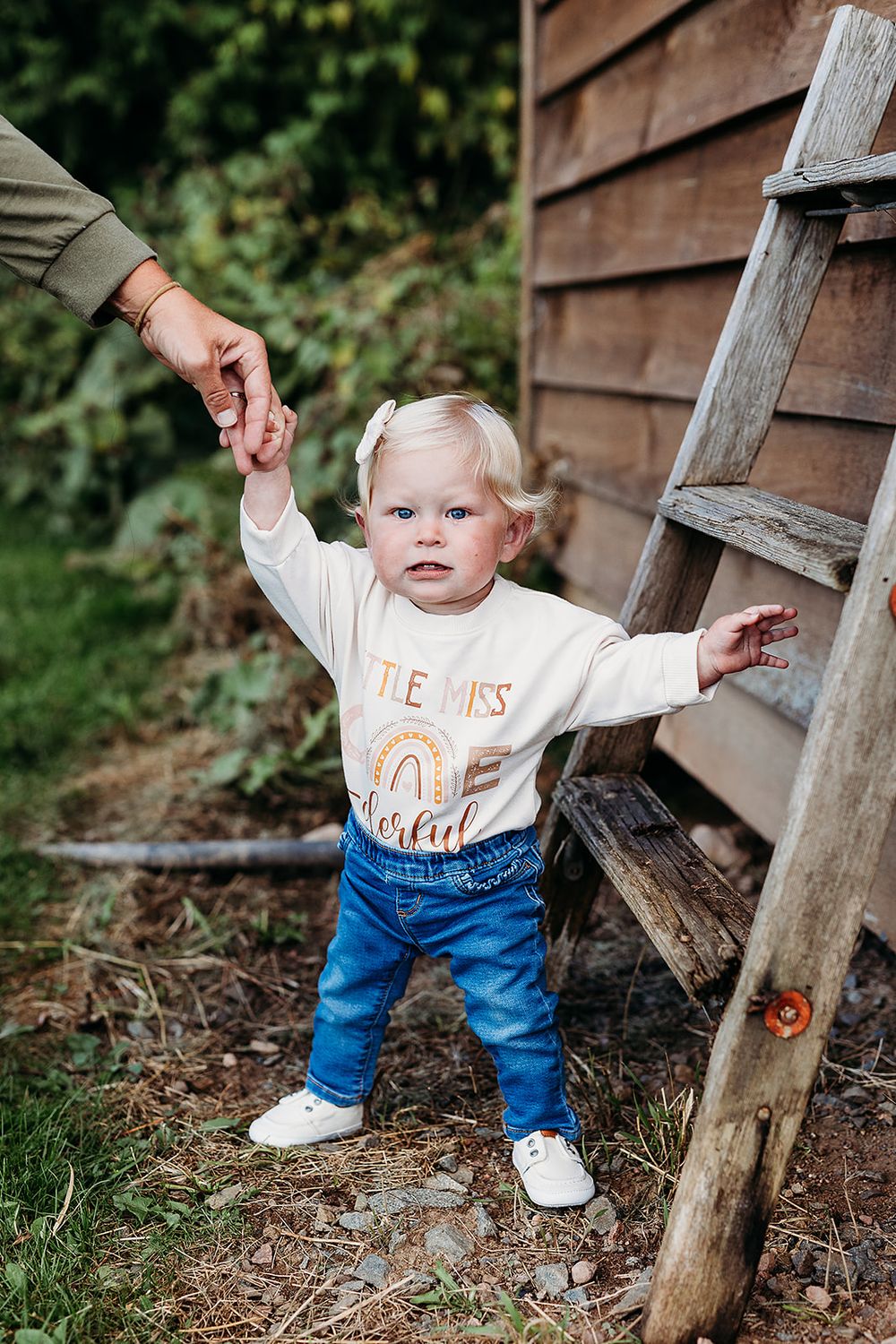 A small child in jeans and white shirt stands next to a wooden ladder while holding an adult's hand for support.
