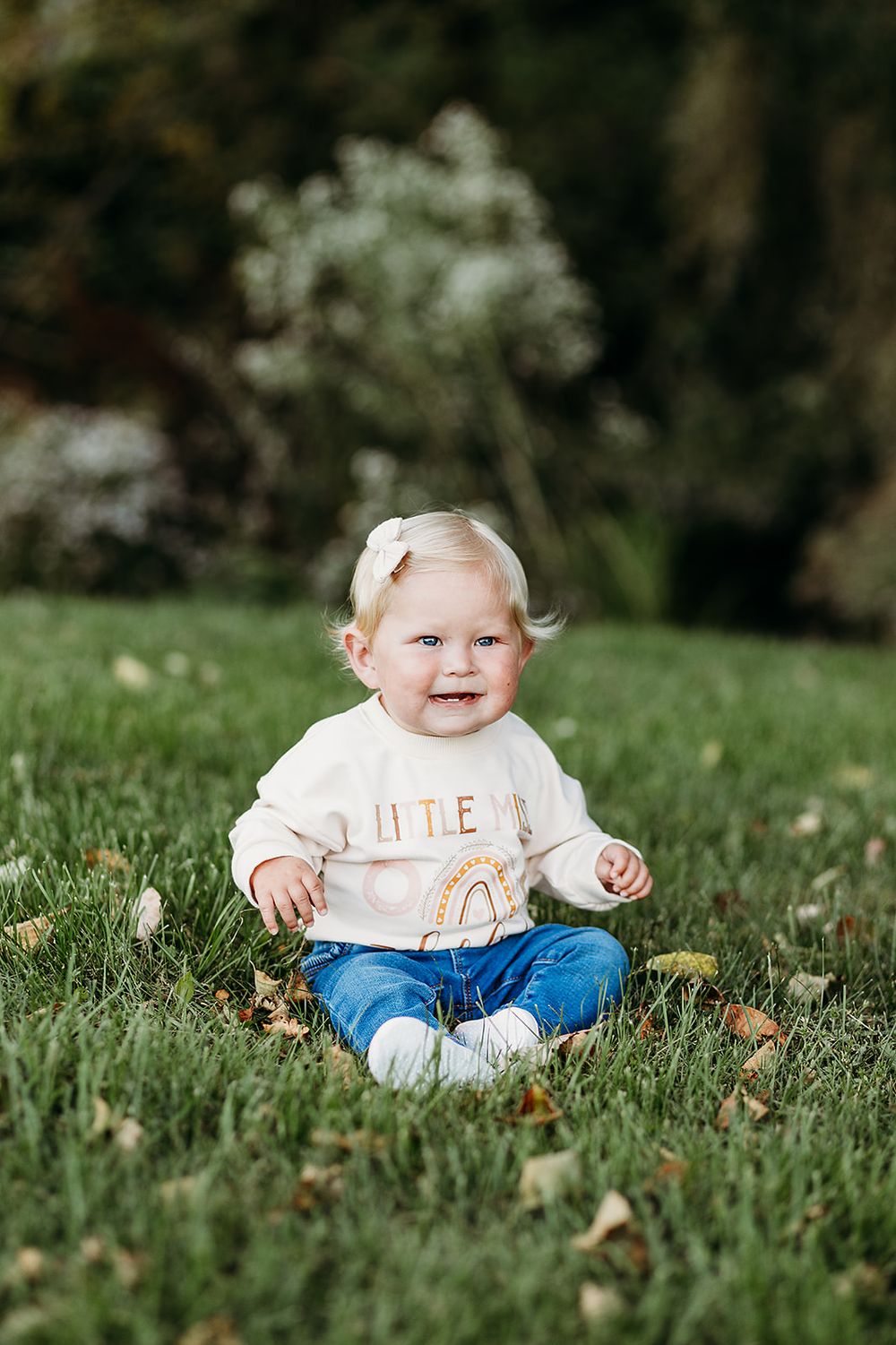 Small child in white sweater and jeans sits on grassy lawn among scattered autumn leaves.