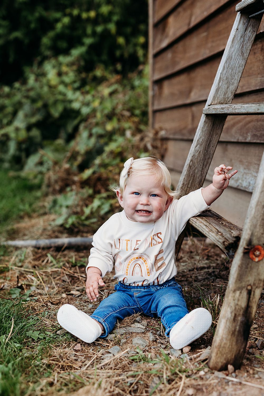 Smiling toddler in jeans and beige top sits beside wooden ladder against rustic barn wall outdoors.