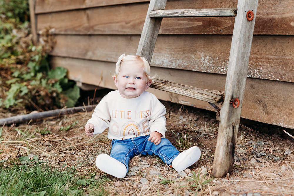 A small child in blue jeans and white sneakers sits on mulch beside an old wooden ladder and barn siding.