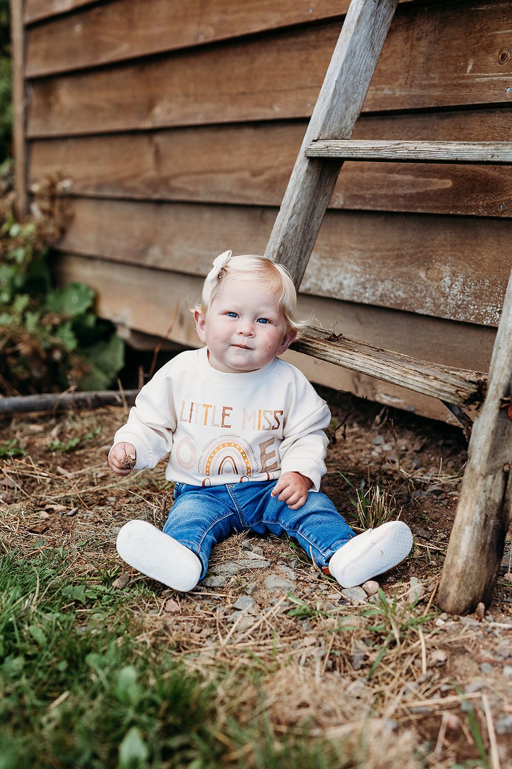 A toddler wearing a cream sweatshirt and jeans sits on straw beside a rustic wooden wall.