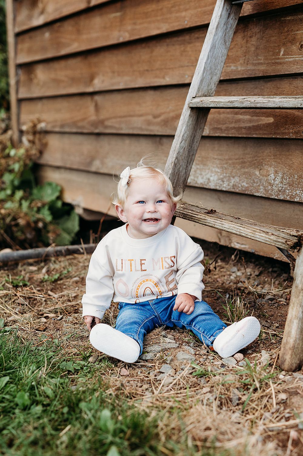A young child sits against a wooden wall wearing a white sweater, blue jeans and white shoes in an outdoor setting.
