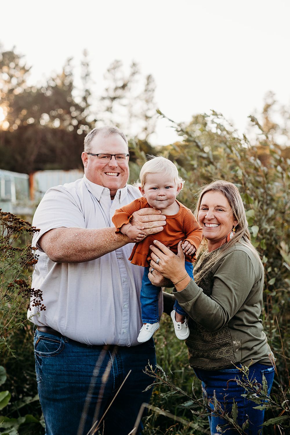 A joyful outdoor family photo shows three people embracing and smiling together at sunset among garden greenery.