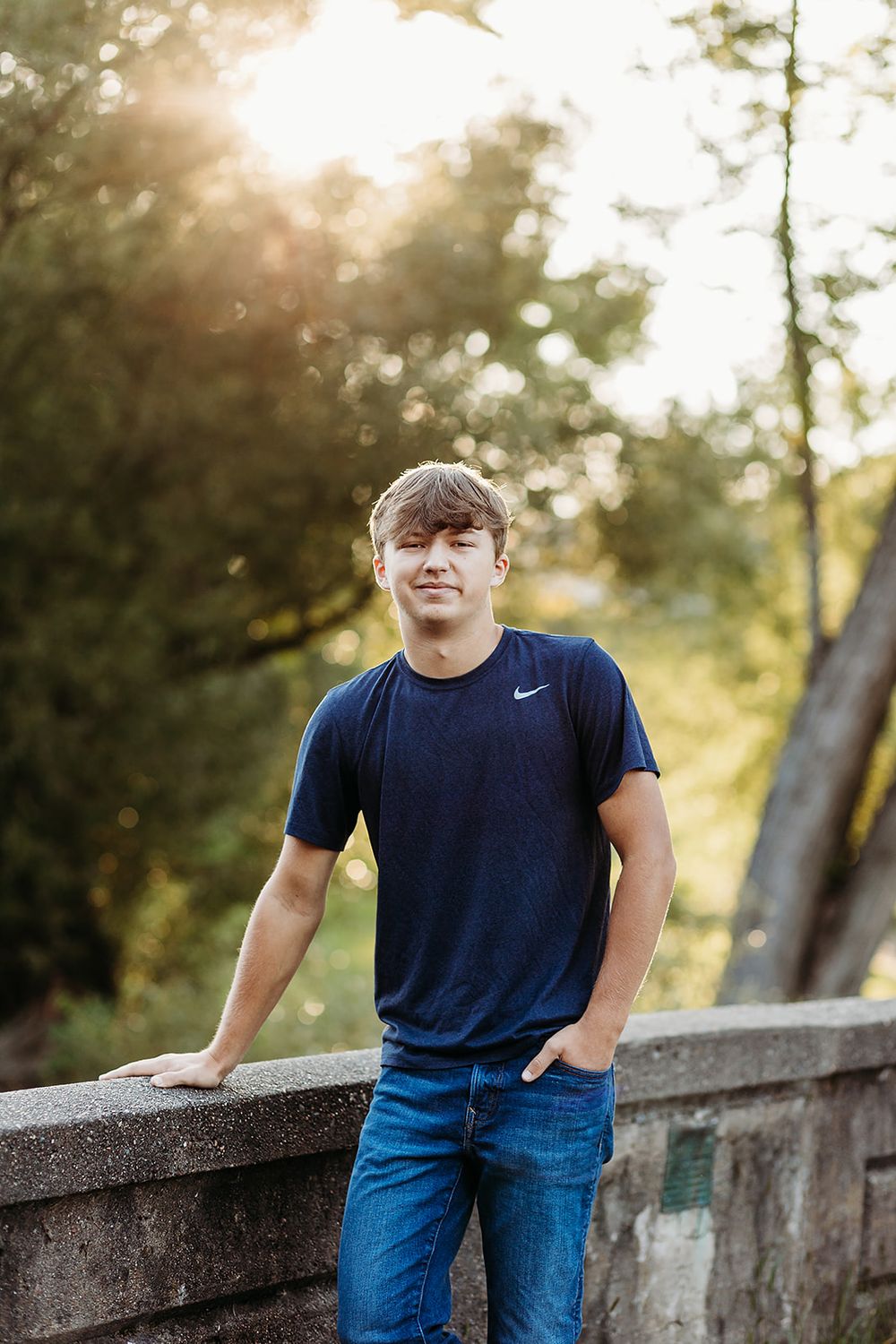 A young man wearing a navy Nike t-shirt and jeans poses casually against a concrete wall in warm sunset lighting.