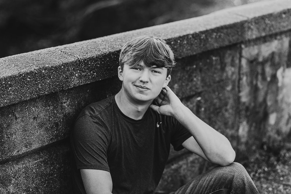 Black and white portrait of a person in casual t-shirt sitting outdoors against stone wall in contemplative pose.