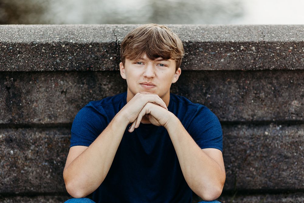 A person in a navy blue polo shirt sits thoughtfully with their chin resting on clasped hands against a stone wall.