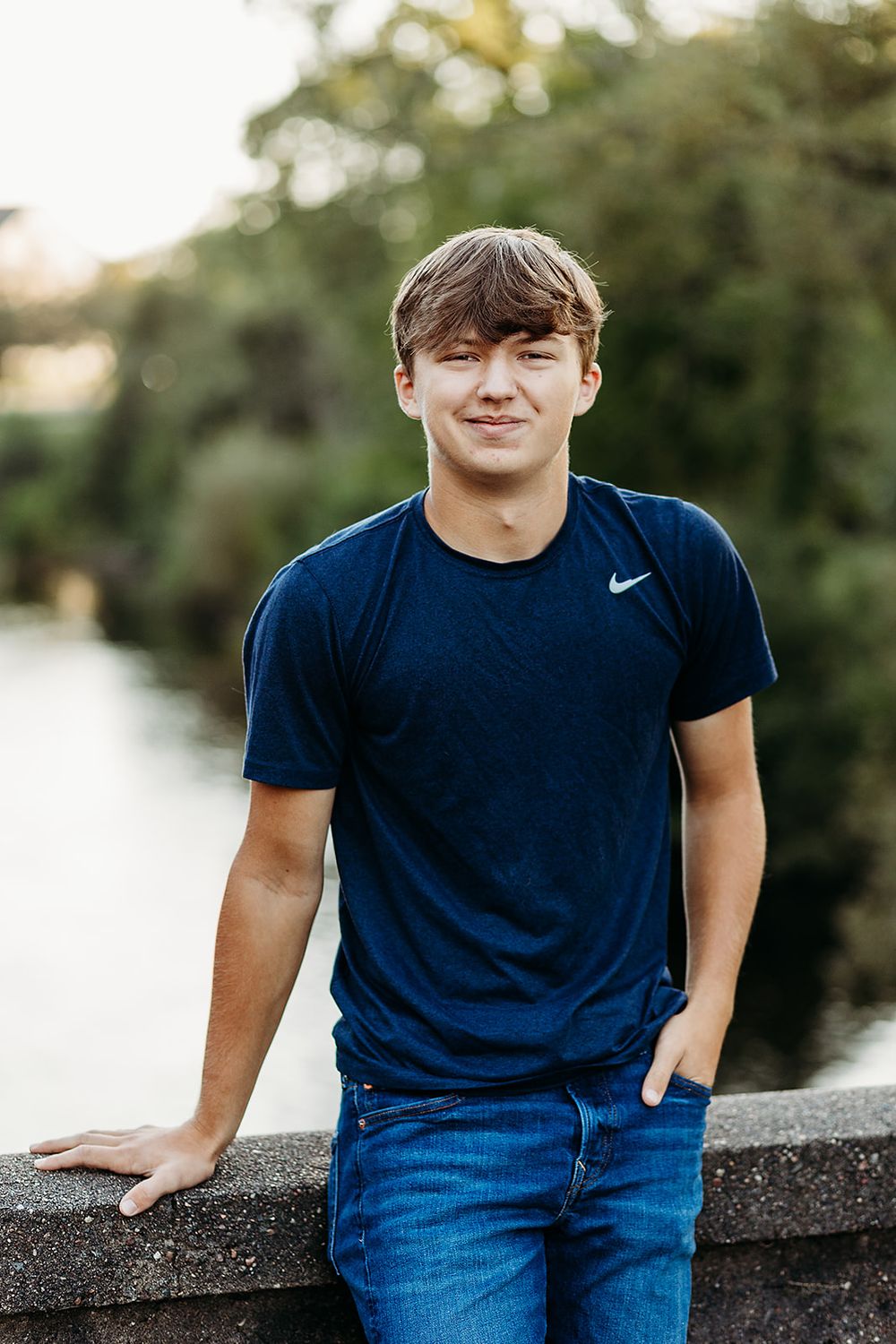 A young person in a navy Nike t-shirt and jeans leans casually against a stone wall near a waterway at sunset.