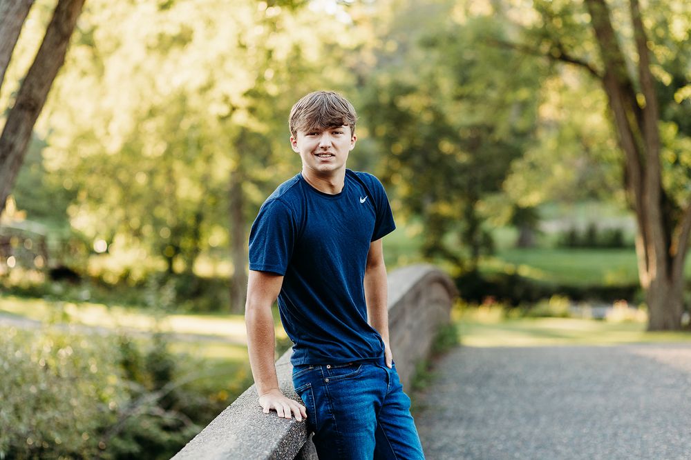 Someone in a navy blue t-shirt and jeans stands casually on a path in a sunlit park during golden hour.