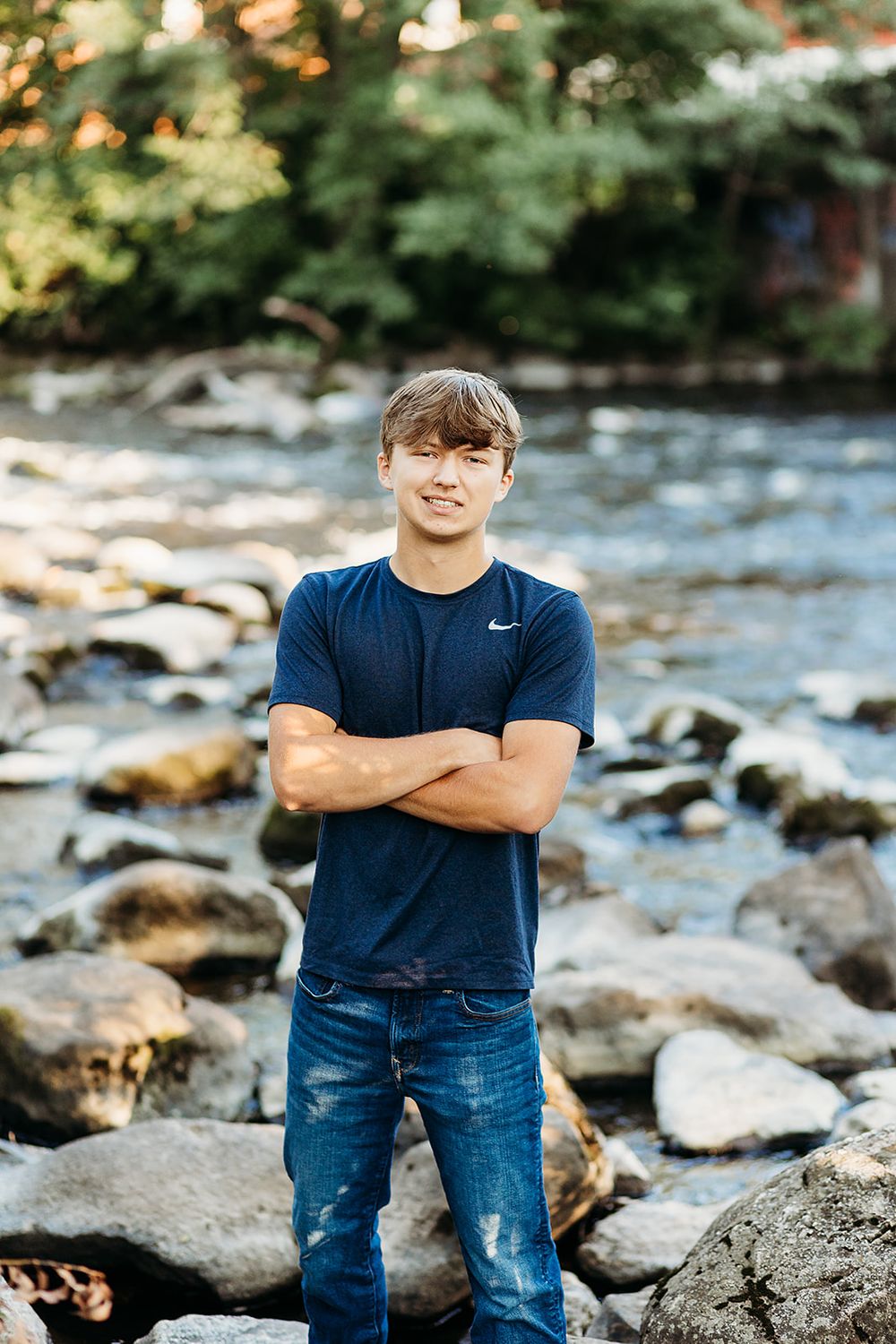 Person in a navy Nike t-shirt and jeans stands with crossed arms on rocky riverbank with flowing water and trees behind.