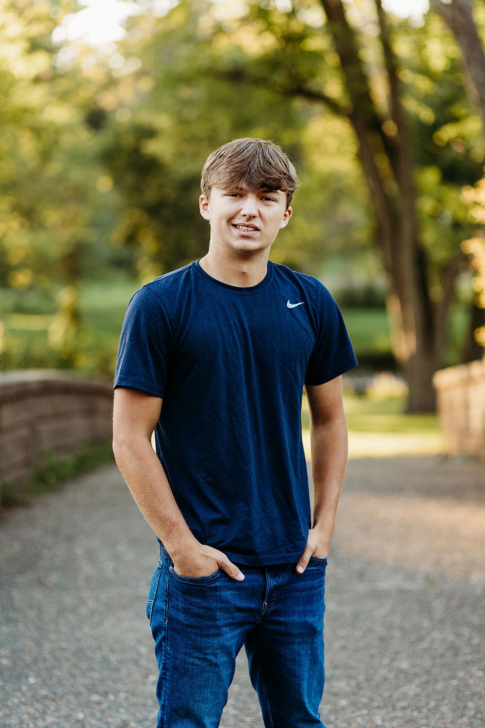 Portrait of someone in a navy Nike t-shirt and jeans standing on a path with trees in soft focus background.