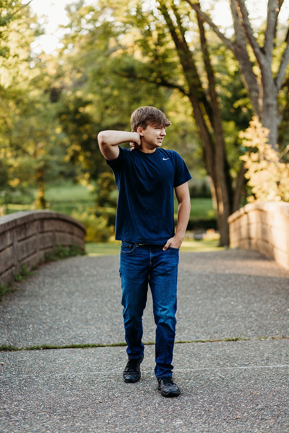 Young person in navy t-shirt and jeans poses casually on gravel path with trees and sunlight in background.