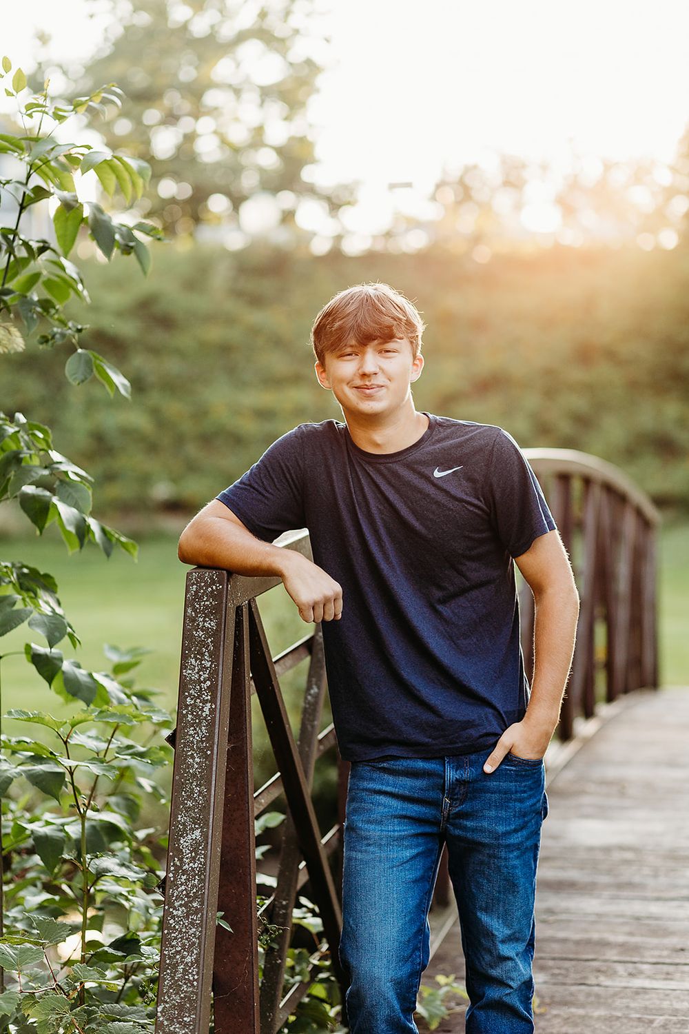 A young person in a Nike t-shirt and jeans poses casually on a wooden footbridge during golden hour.