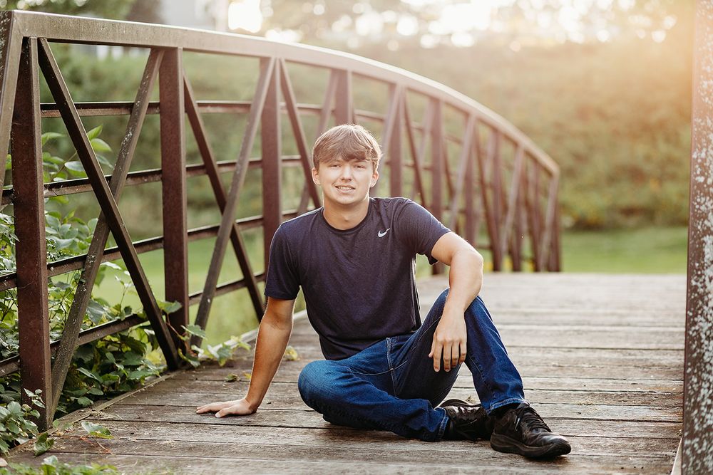 Young person in navy t-shirt and jeans sits casually on rustic wooden bridge during golden hour sunset.