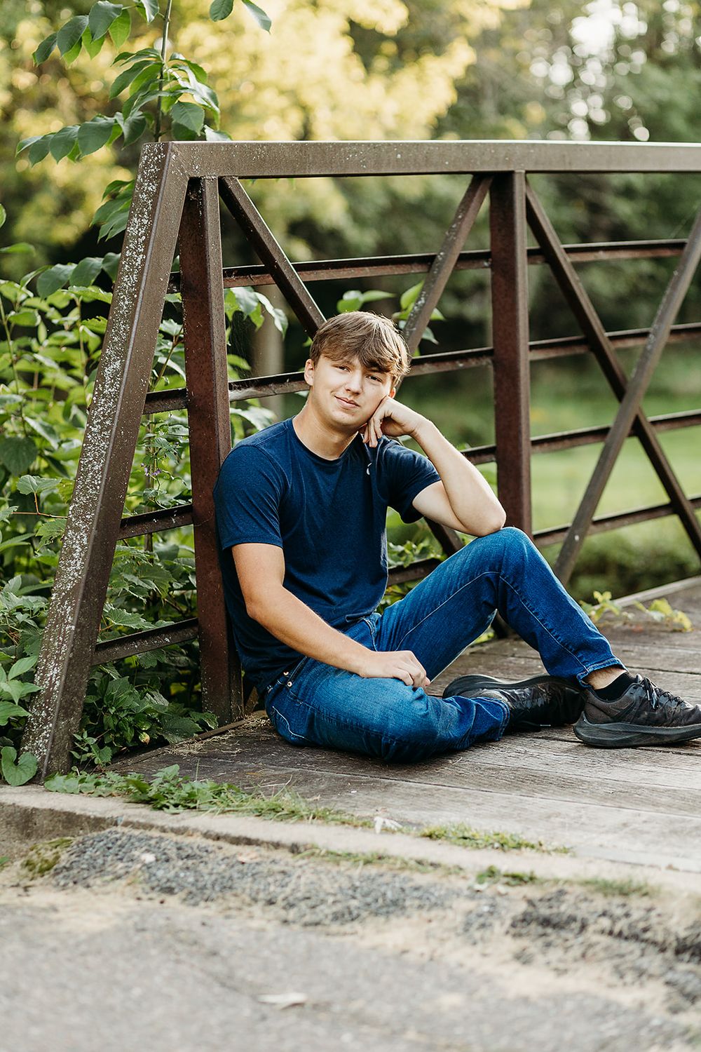 A casual outdoor portrait of someone sitting relaxed on a wooden bridge railing wearing a navy shirt and jeans.