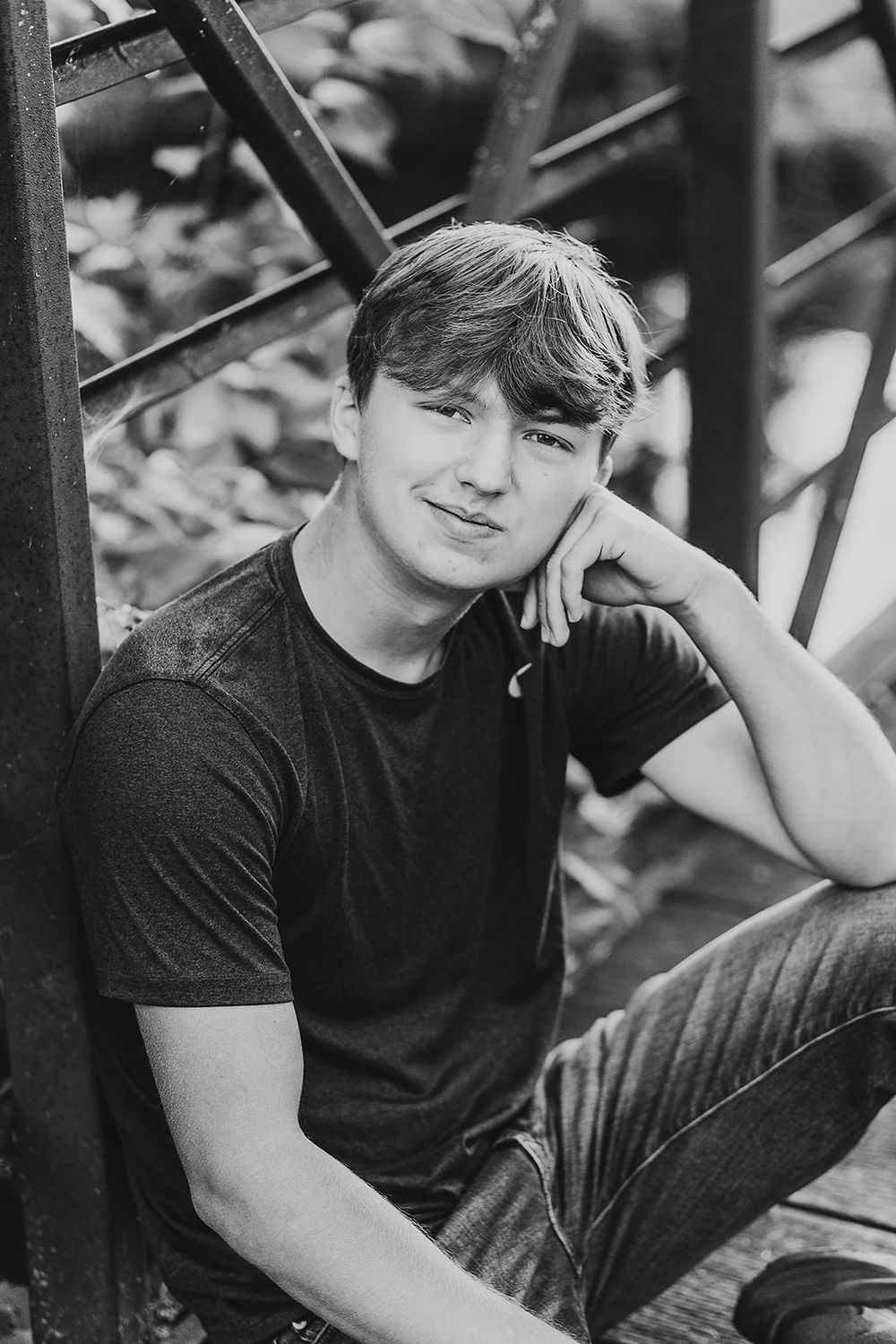 Black and white portrait of young person in casual t-shirt leaning against metal structure outdoors.