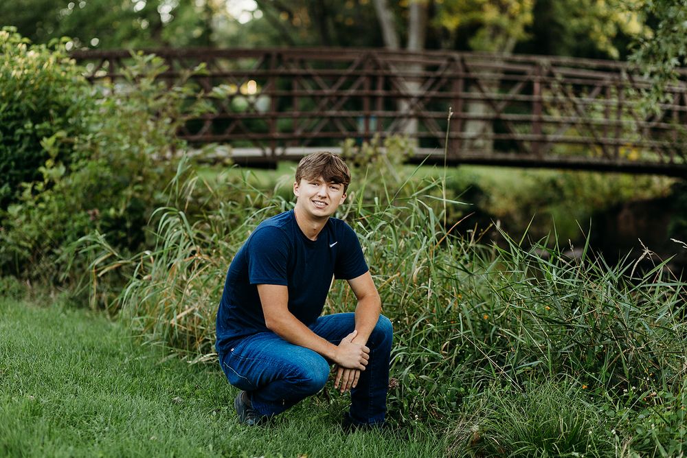A person in blue casual attire kneels on green grass near a rustic bridge surrounded by lush vegetation.