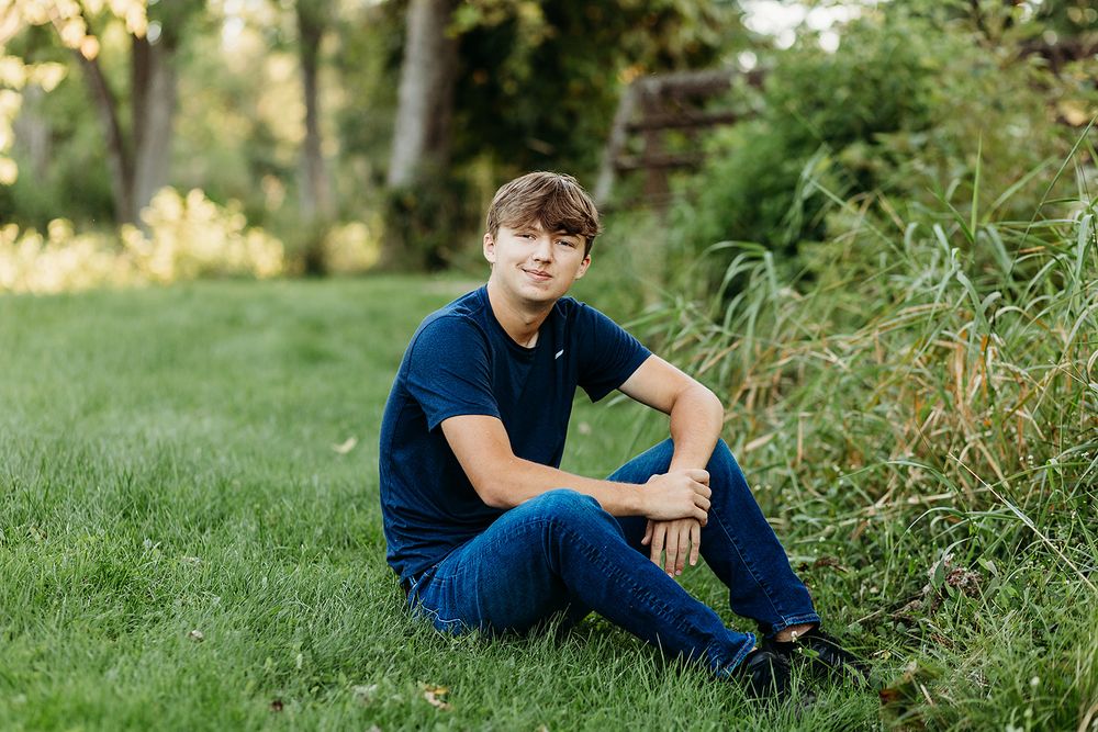 A young person in a navy shirt and jeans sits on grass near bushes in a garden setting during golden hour.