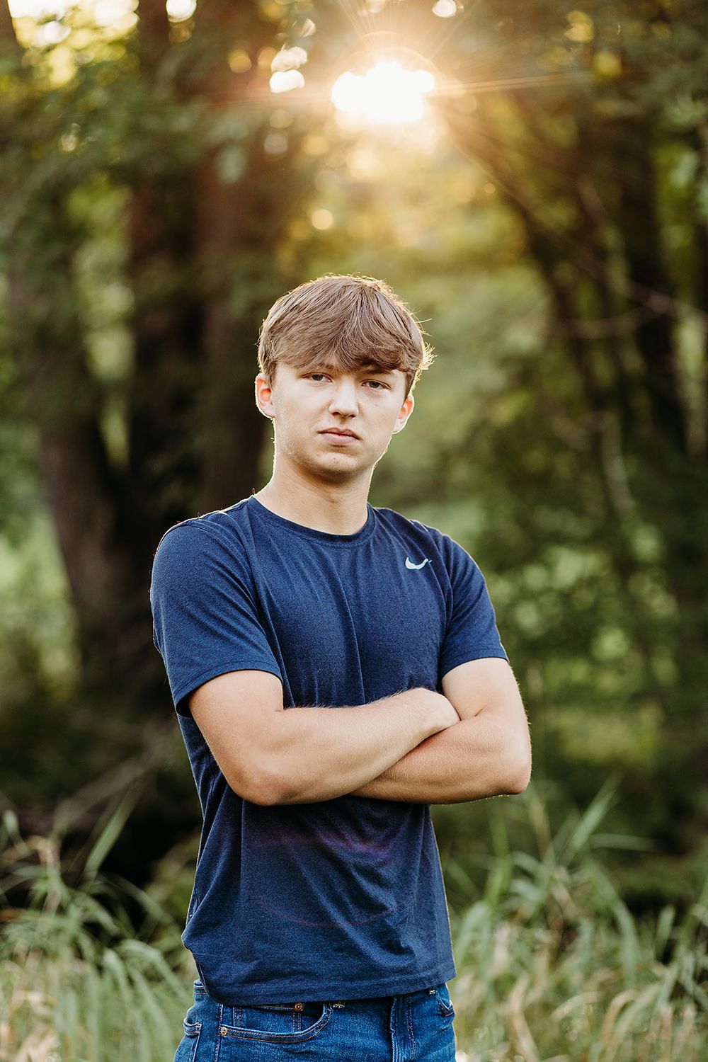 A figure stands with crossed arms wearing a navy Nike t-shirt in a sunlit wooded area during golden hour.