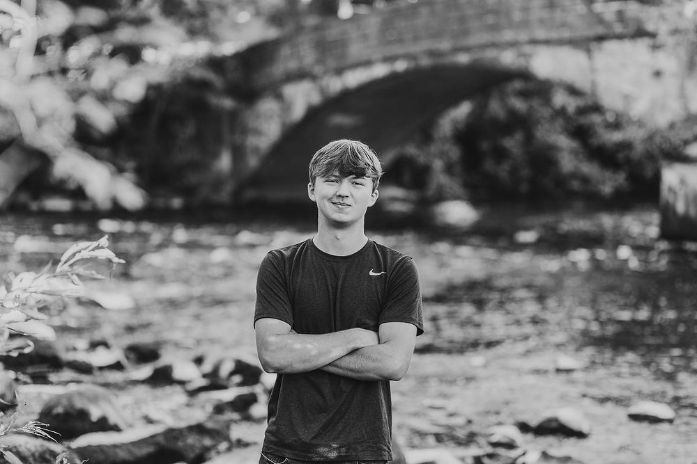 A black and white portrait showing a person in a Nike shirt standing in front of a stone bridge over a rocky stream.