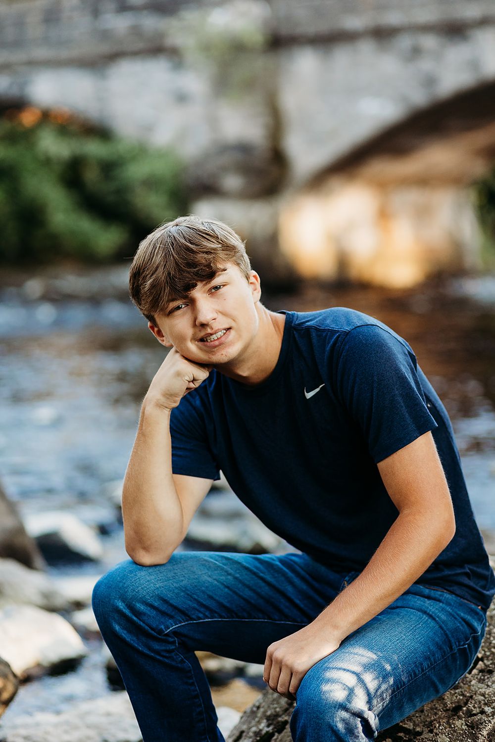 Person in a navy Nike t-shirt and jeans poses on rocks near a stream in an outdoor portrait setting.