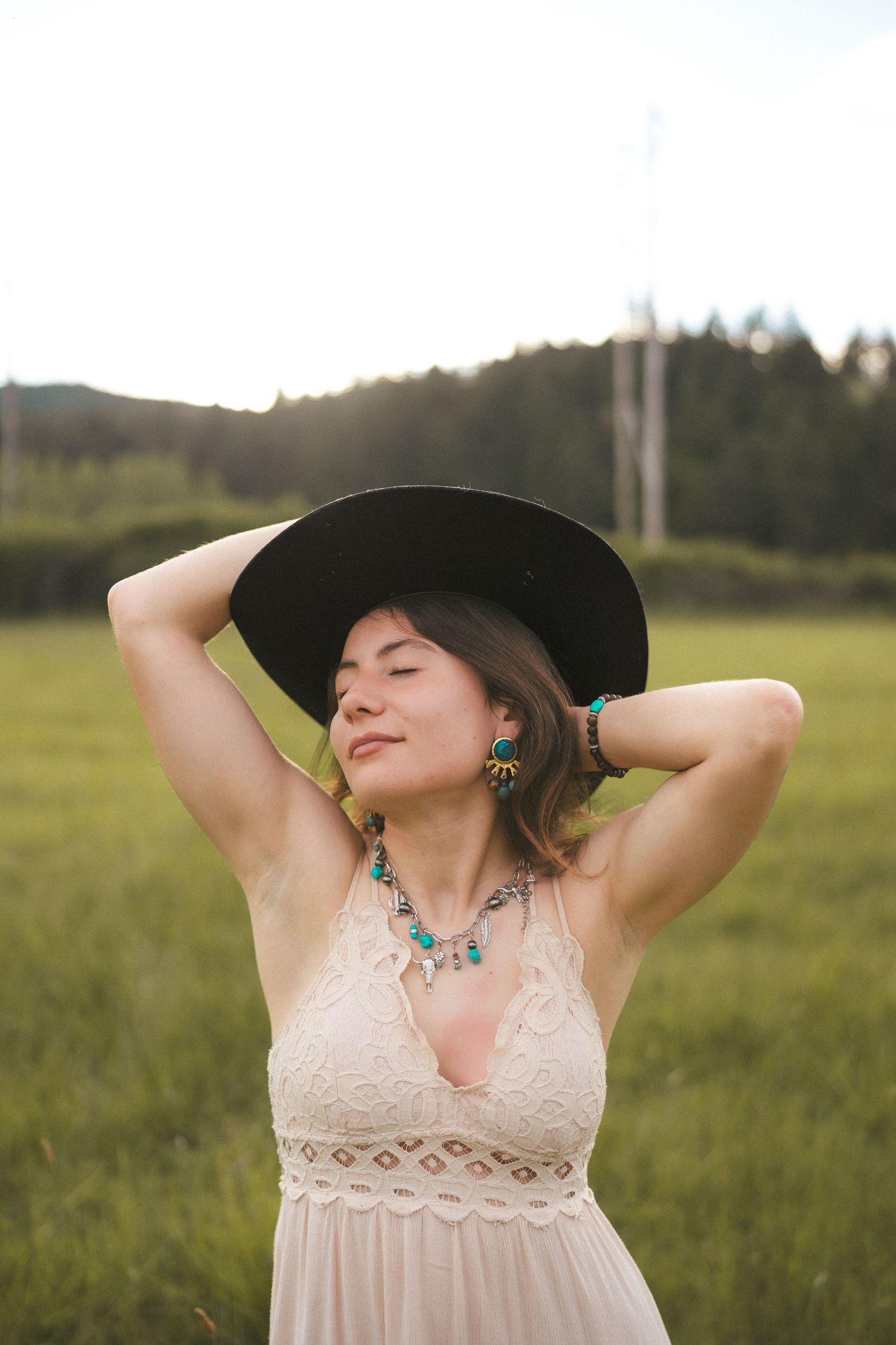 A person in a white lace dress and black hat poses in a sunlit field.